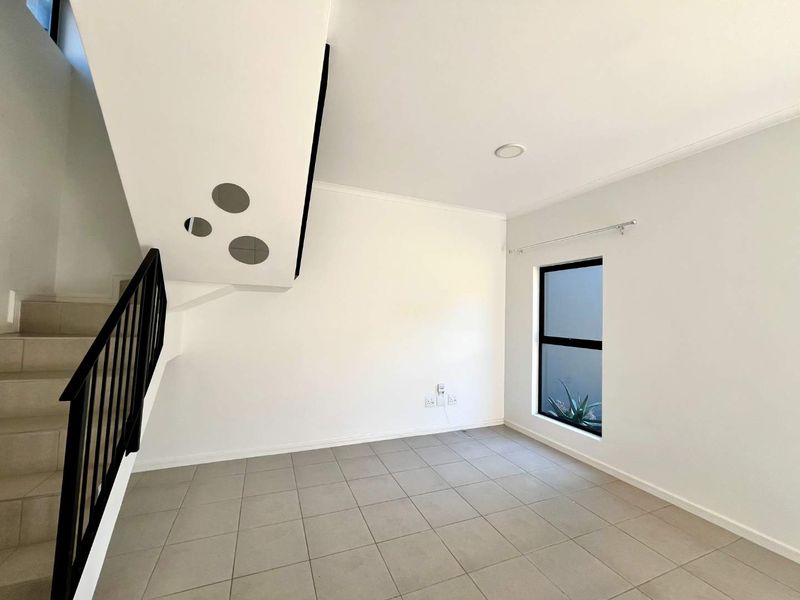 A bright, open-plan living area featuring neutral tiled flooring, a modern staircase with a black metal balustrade, and a large window providing natural light.