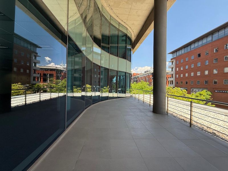 A modern, curved balcony featuring tiled flooring, a stainless steel railing, and a prominent concrete support column, offering views of the surrounding office complex.