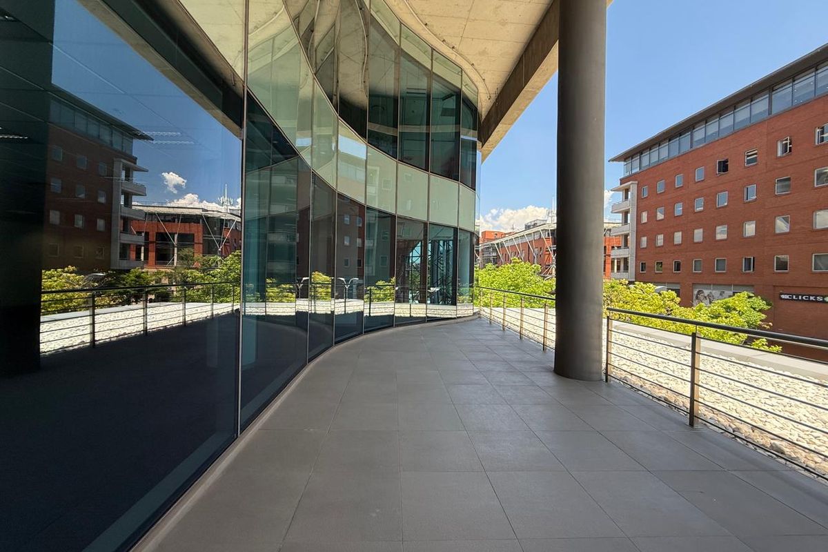 A modern, curved balcony featuring tiled flooring, a stainless steel railing, and a prominent concrete support column, offering views of the surrounding office complex.