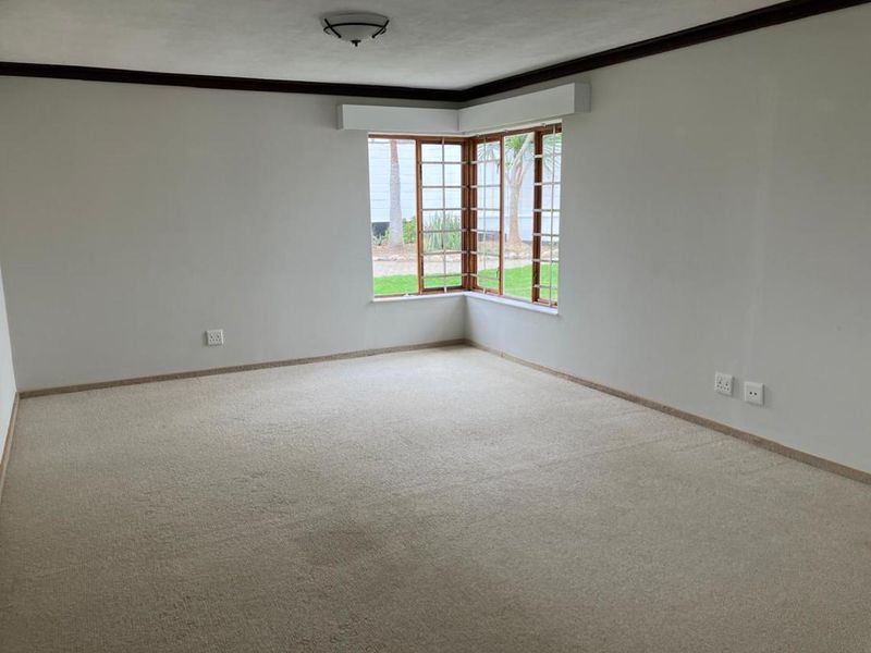 A spacious, empty living room featuring neutral carpeting, crown molding, and a large bay window that provides natural light and garden views.