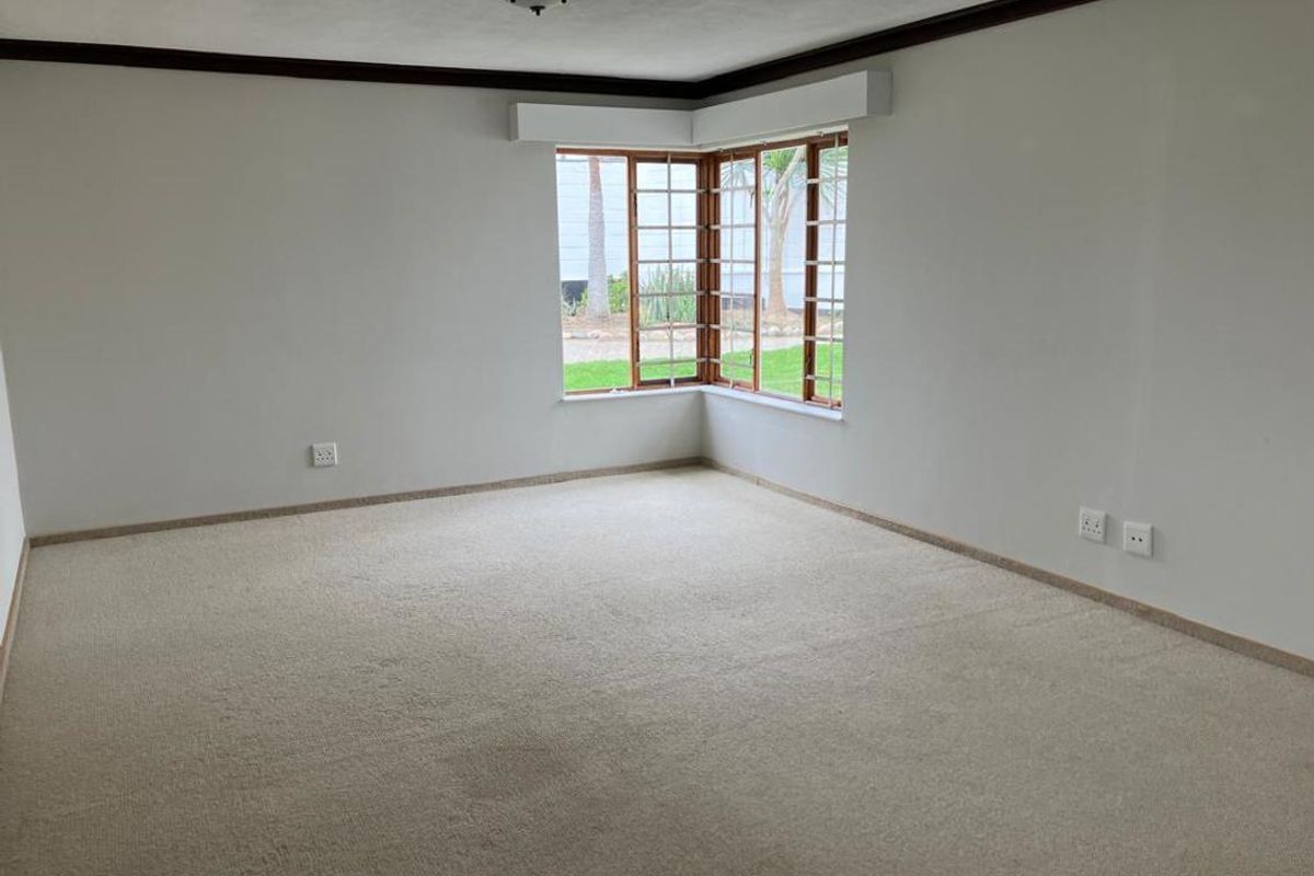 A spacious, empty living room featuring neutral carpeting, crown molding, and a large bay window that provides natural light and garden views.