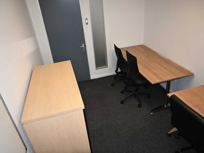 A functional office space featuring carpeted flooring, a solid core door, and a vertical sidelight window, providing a clean and professional work environment.