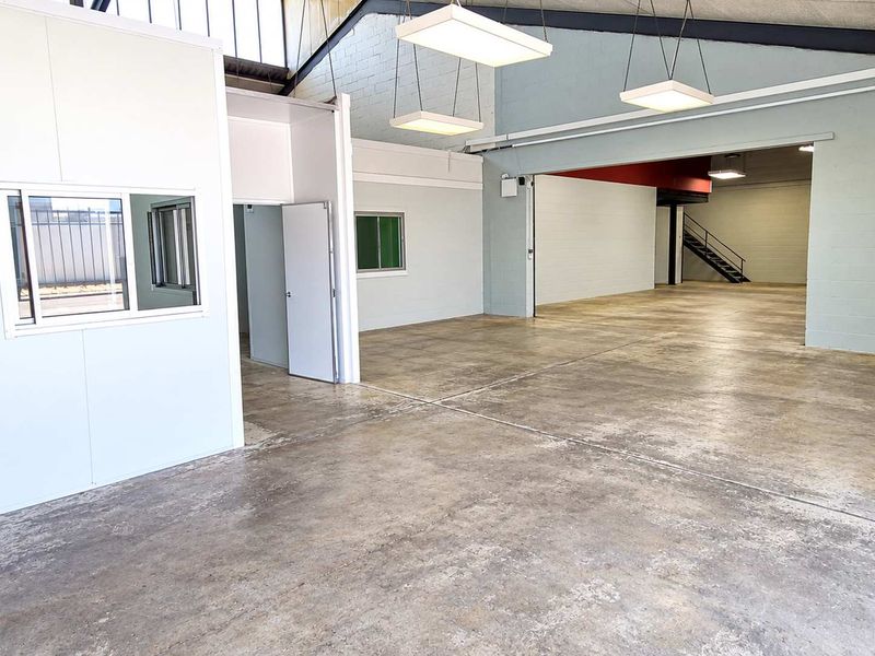 Spacious industrial warehouse area featuring polished concrete floors, high ceilings with exposed trusses, and a partitioned office enclosure with an internal staircase.