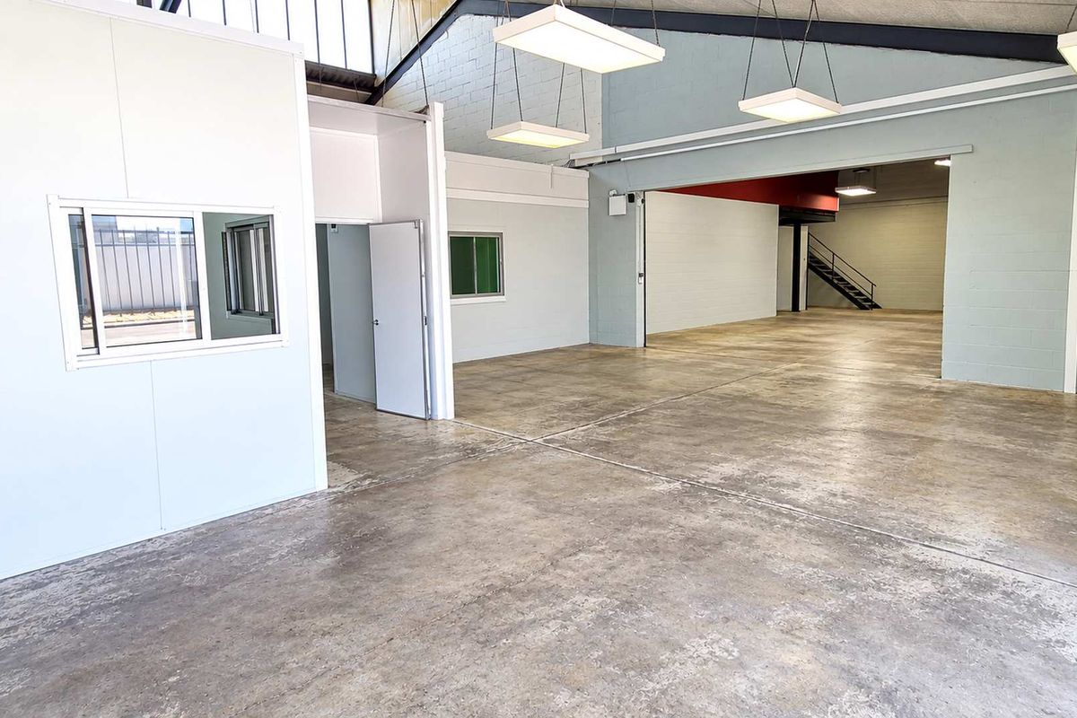 Spacious industrial warehouse area featuring polished concrete floors, high ceilings with exposed trusses, and a partitioned office enclosure with an internal staircase.