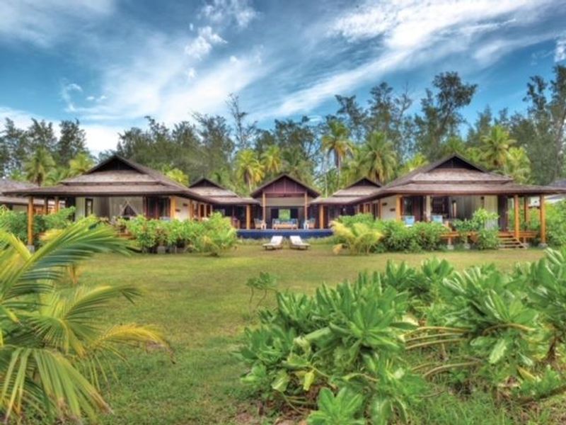 A scenic rear view of a villa featuring traditional pitched roofs, expansive wooden decking, and lush tropical landscaping surrounding the property.