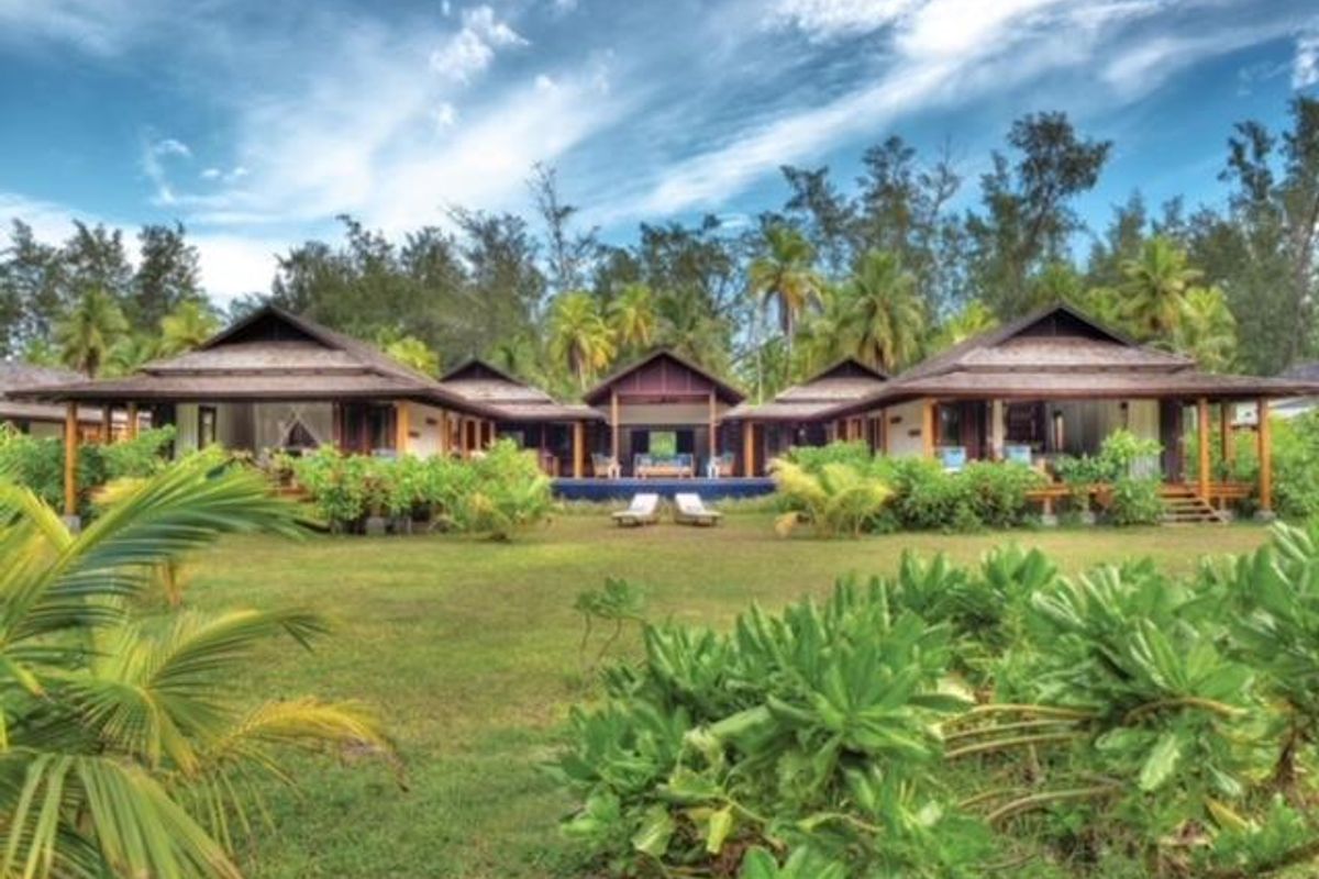 A scenic rear view of a villa featuring traditional pitched roofs, expansive wooden decking, and lush tropical landscaping surrounding the property.
