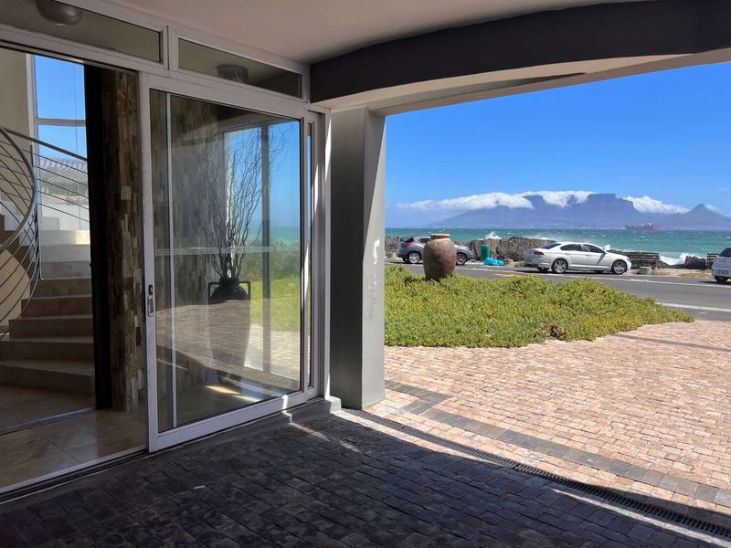 A covered patio area featuring stone cladding, paved flooring, and sliding glass doors, offering a direct view of the coastal road and Table Mountain.