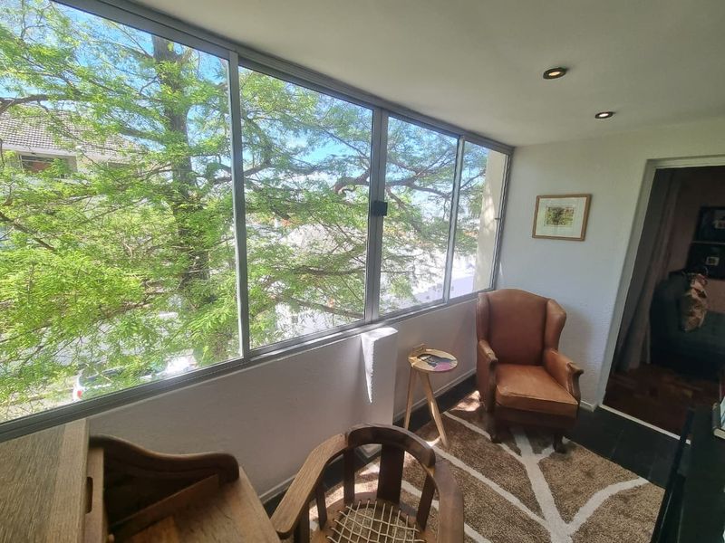 A bright, enclosed balcony featuring large windows with leafy views, recessed lighting, and tiled flooring, offering a versatile space for relaxation.