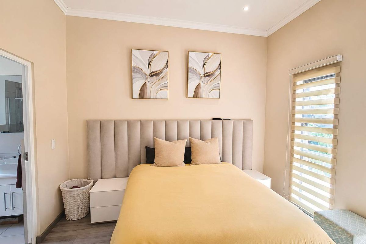 A well-maintained bedroom featuring a modern upholstered headboard, neutral wall tones, and a window with fitted roller blinds, leading into an en-suite bathroom.