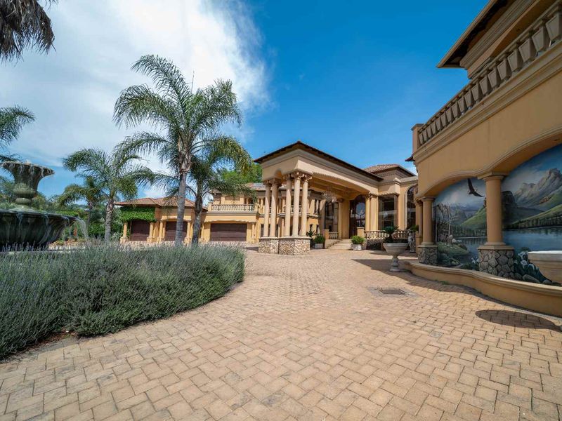 Grand front entrance of a small holding featuring a paved driveway, stone-clad columns, a decorative fountain, and a large exterior mural.