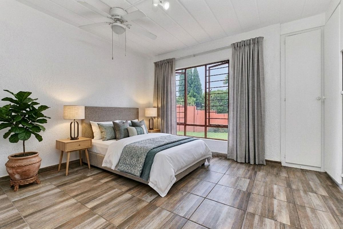 A well-lit bedroom featuring wood-look tile flooring, a ceiling fan, and a large window with security bars overlooking the garden.