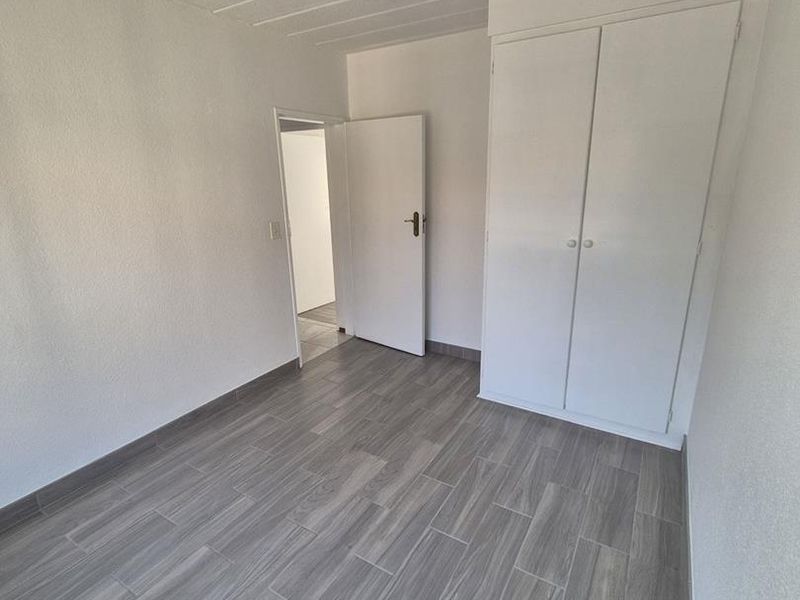A clean, empty bedroom featuring modern grey wood-look floor tiles, a built-in wardrobe, and a neutral white finish throughout.
