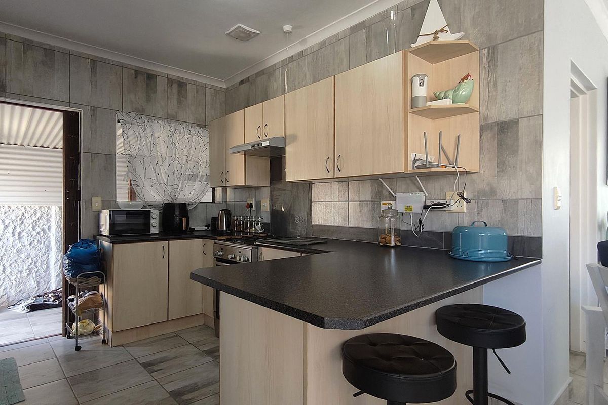 A functional kitchen featuring light wood cabinetry, dark laminate countertops, and grey tiled walls, with an integrated breakfast bar and direct access to the patio.