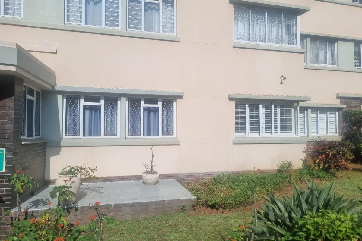 Rear exterior view of the apartment building featuring a concrete patio, landscaped garden, and windows fitted with security bars.