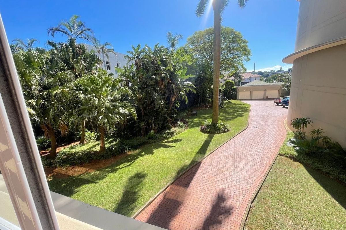 An elevated view of a well-maintained garden area featuring a brick-paved driveway leading to a garage block, surrounded by lush tropical landscaping.