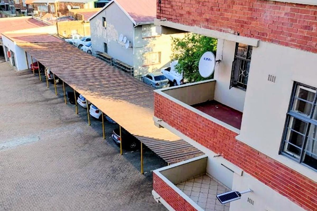 Elevated view of the apartment building exterior, featuring brick balconies, a covered carport area, and paved parking facilities.