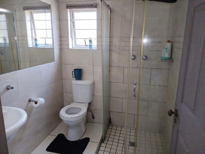 A functional bathroom featuring a glass shower enclosure, wall-mounted basin, toilet, and neutral wall tiling with natural light from a window.