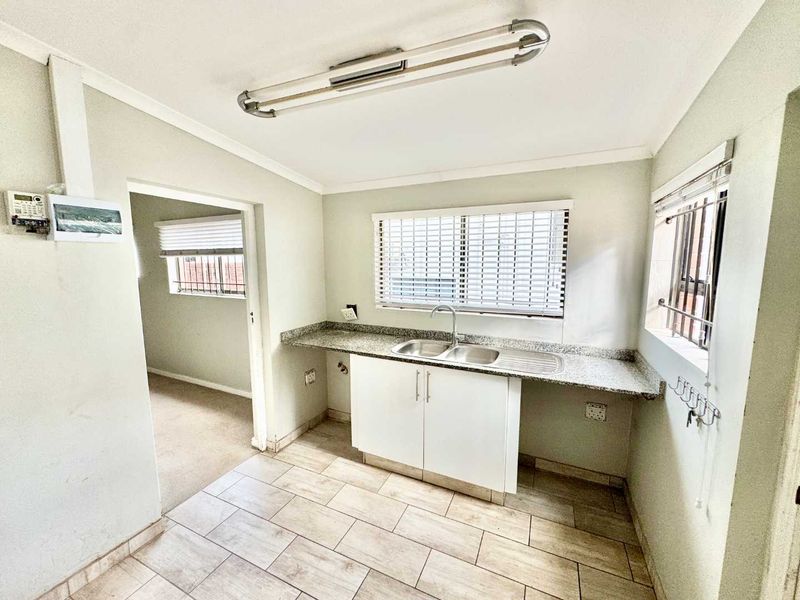 A functional scullery area featuring granite countertops, a double stainless steel sink, tiled flooring, and ample natural light through fitted venetian blinds.