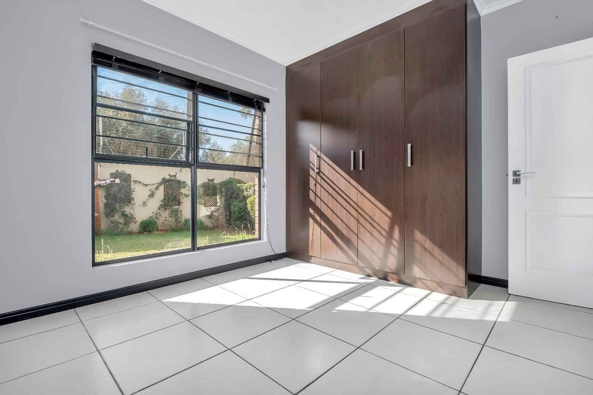 A bright bedroom featuring large white floor tiles, a dark wood built-in wardrobe, and a window with security bars overlooking the garden.