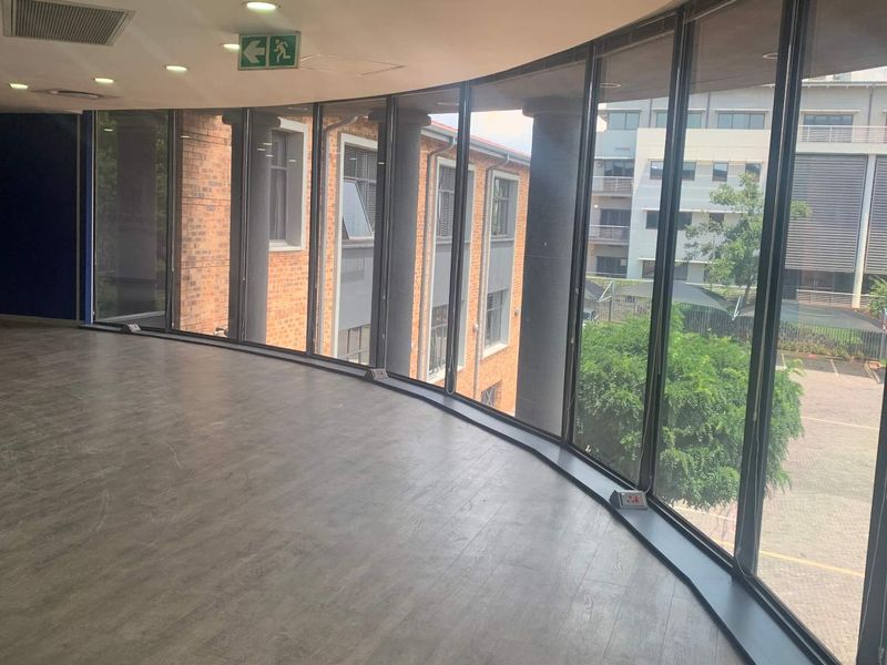 Spacious empty office area featuring large curved floor-to-ceiling windows, recessed lighting, and modern laminate flooring with views of the surrounding office park.