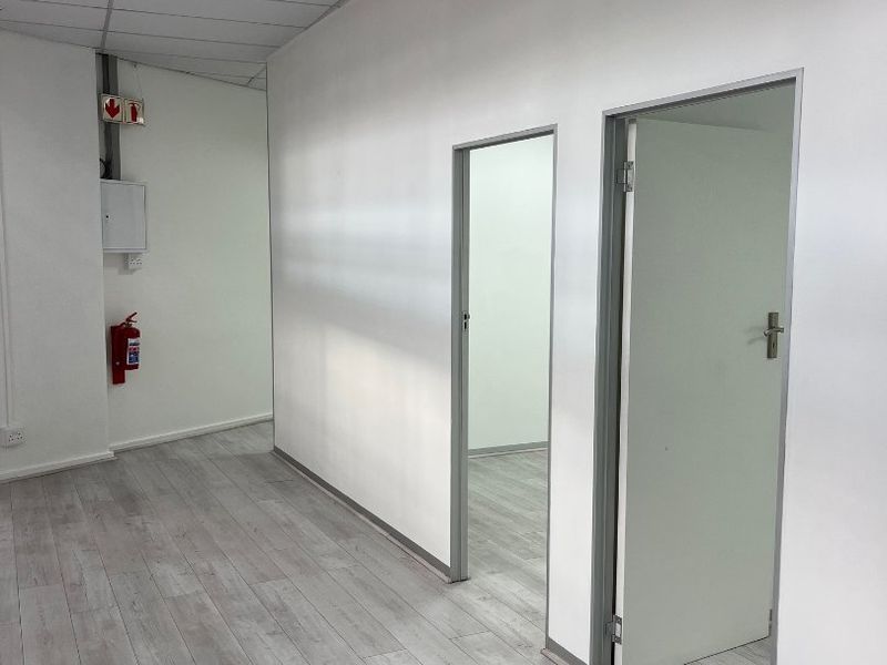 A clean, modern commercial hallway featuring light wood-look laminate flooring, a suspended ceiling, and access to adjacent office or utility rooms.