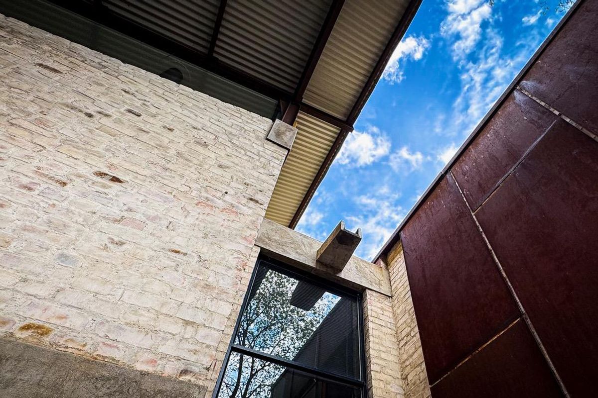 A modern architectural exterior detail featuring a mix of exposed brick, corten steel cladding, and corrugated metal roofing with a large window.