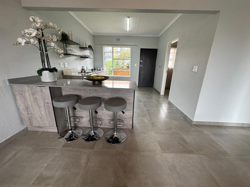 Modern open-plan kitchen featuring a breakfast bar, stone countertops, floating shelving, and neutral tiled flooring, creating a functional and contemporary living space.