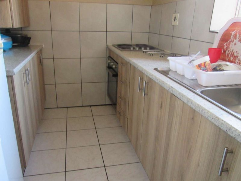 A functional galley-style kitchen featuring light wood-look cabinetry, stone-effect countertops, built-in oven and hob, and neutral ceramic wall and floor tiling.