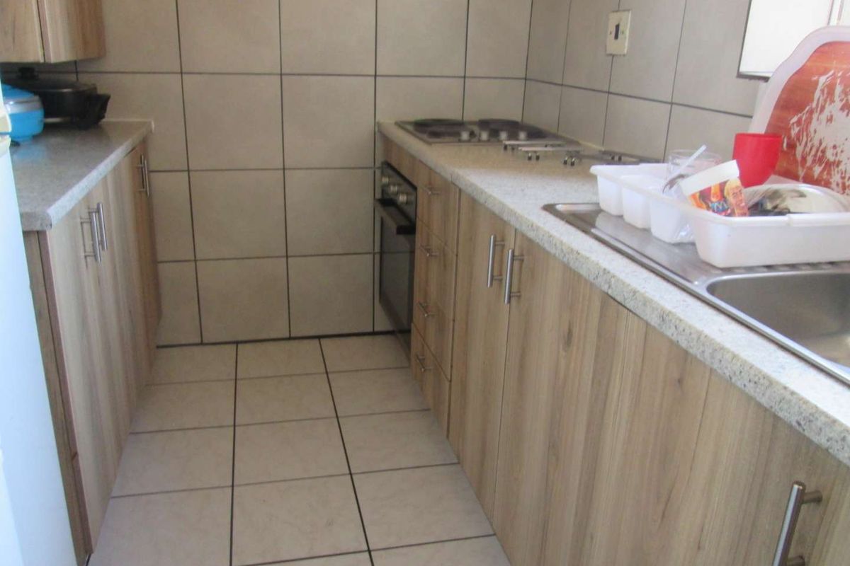 A functional galley-style kitchen featuring light wood-look cabinetry, stone-effect countertops, built-in oven and hob, and neutral ceramic wall and floor tiling.