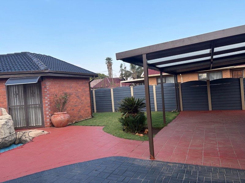 A well-maintained rear exterior featuring a brick cottage facade, a covered carport, paved patio area, and a neat garden with perimeter fencing.