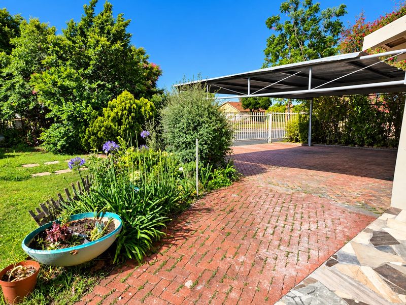 Front exterior view featuring a brick paved driveway, a covered carport, and a well-established garden with a metal entrance gate.