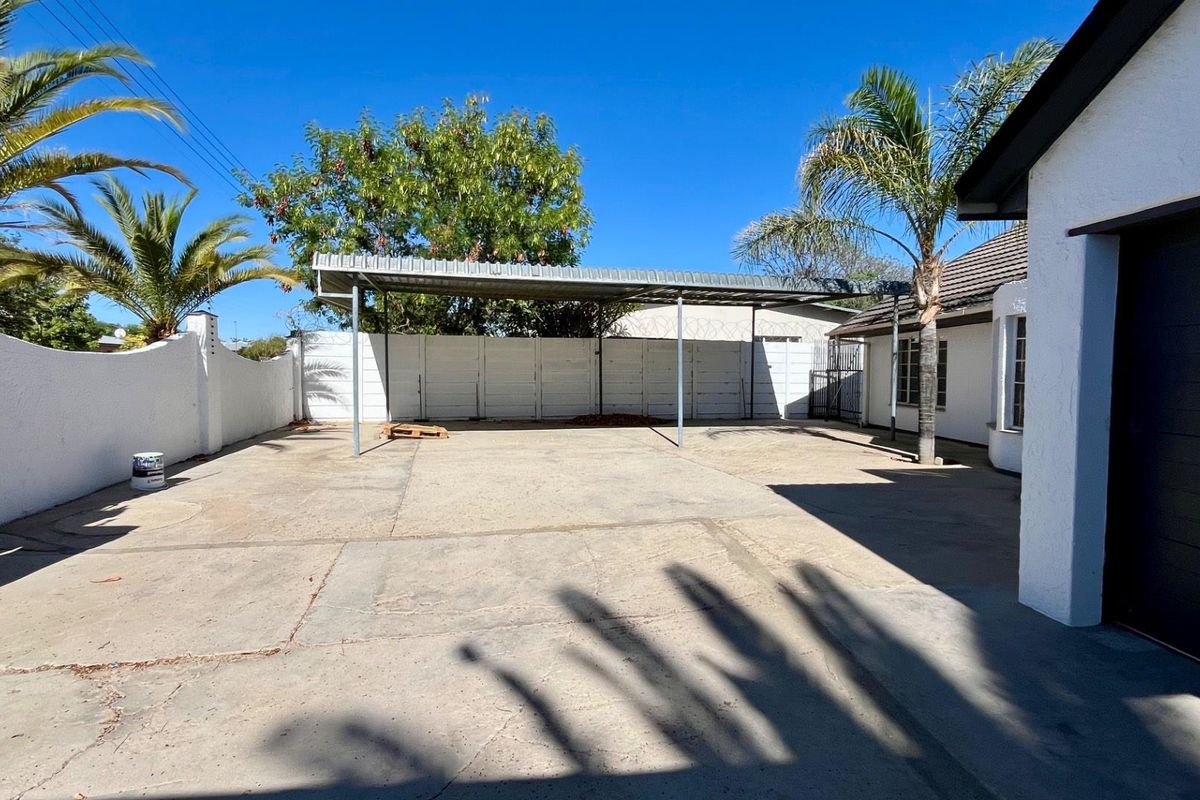 A spacious rear exterior area featuring a concrete driveway, a metal carport structure, and secure perimeter walling.
