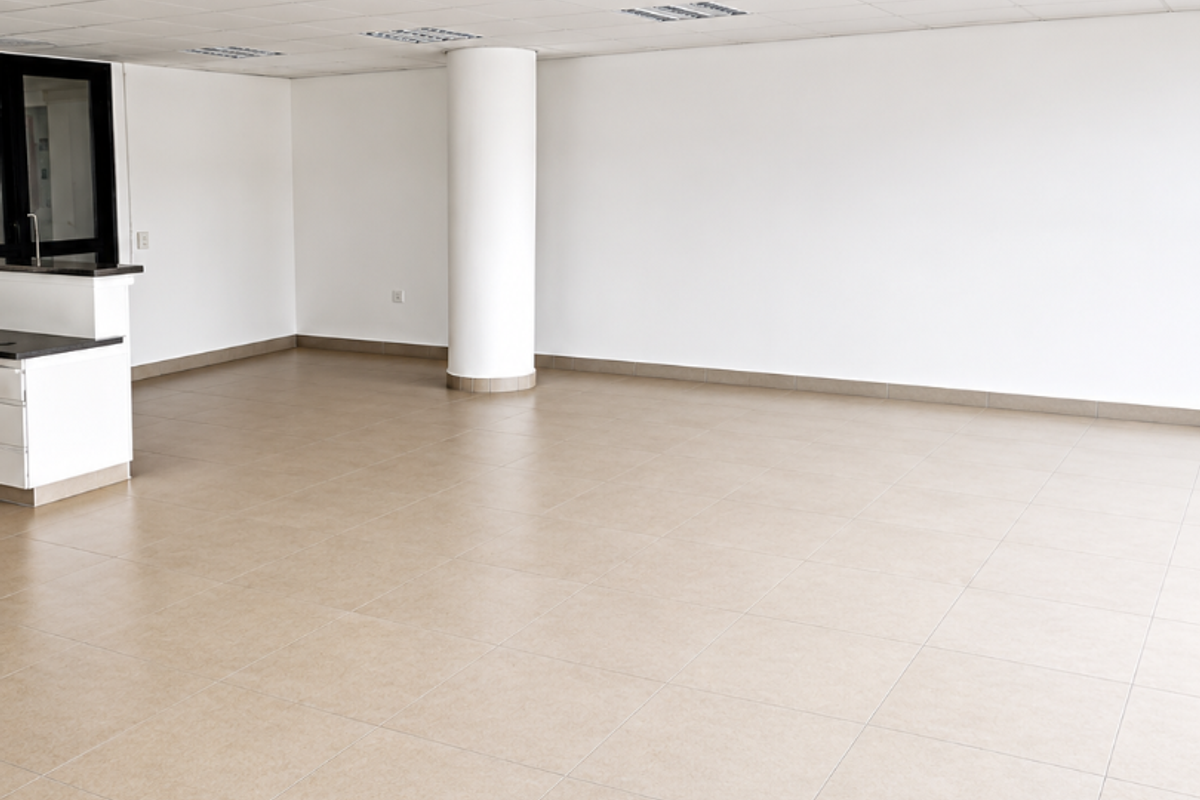 A spacious, open-plan office area featuring neutral ceramic floor tiles, a suspended ceiling with recessed lighting, and a central structural column.