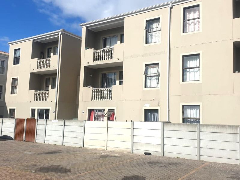 Exterior view of a multi-story apartment complex featuring balconies with balustrades, paved parking, and a secure boundary wall.
