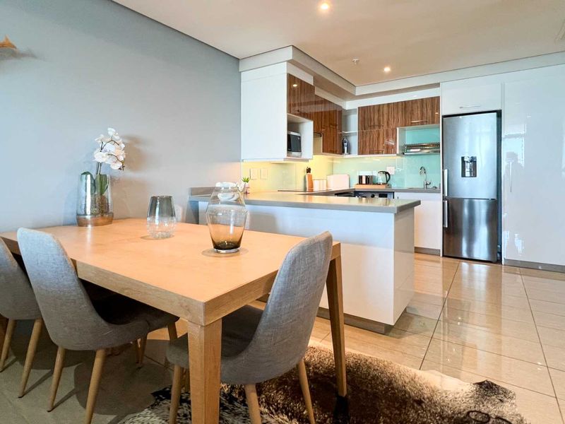Modern open-plan kitchen and dining area featuring sleek cabinetry, stone countertops, integrated appliances, and polished tiled flooring.