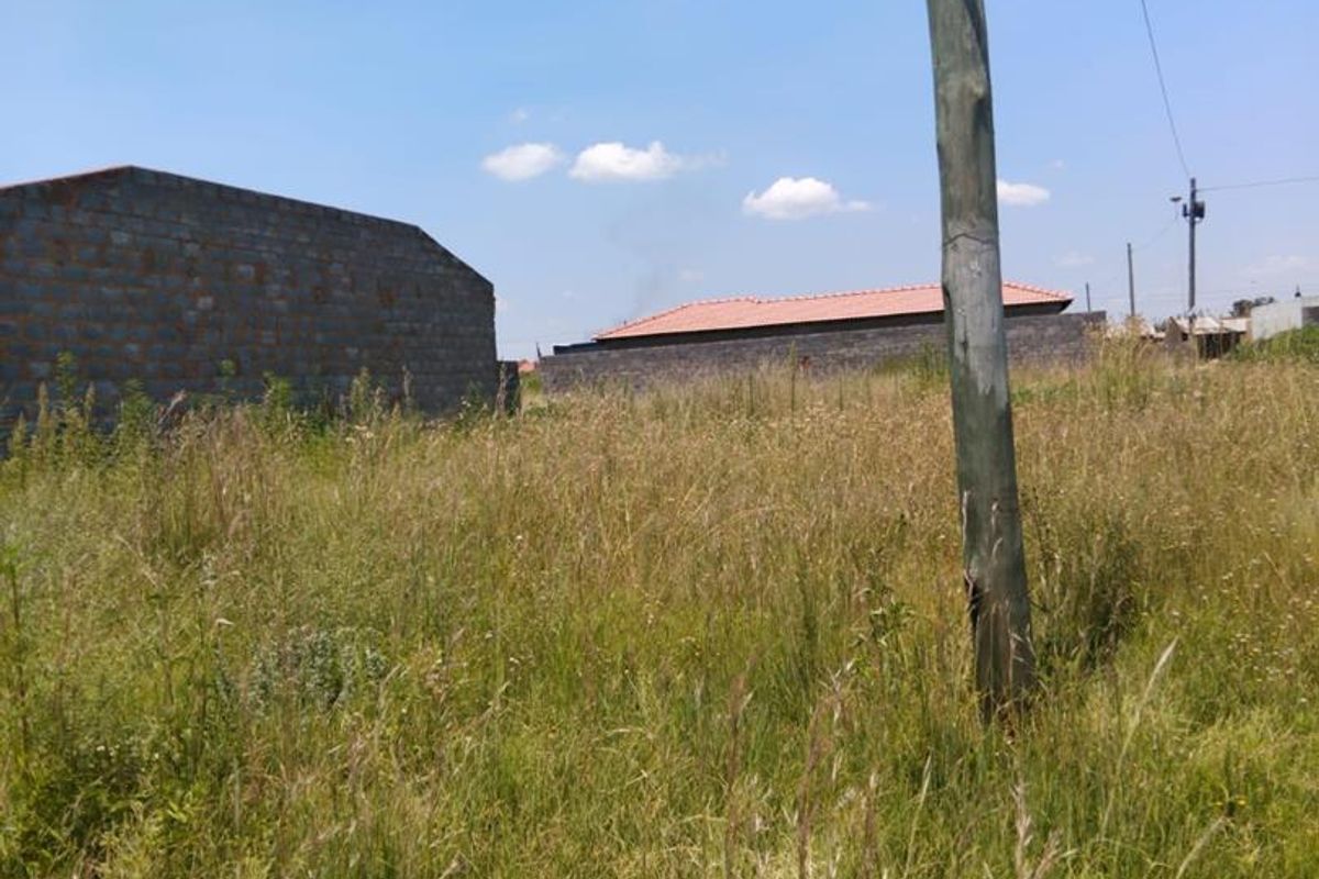 A vacant residential plot featuring tall grass, a utility pole, and surrounding boundary walls, offering a blank canvas for development.