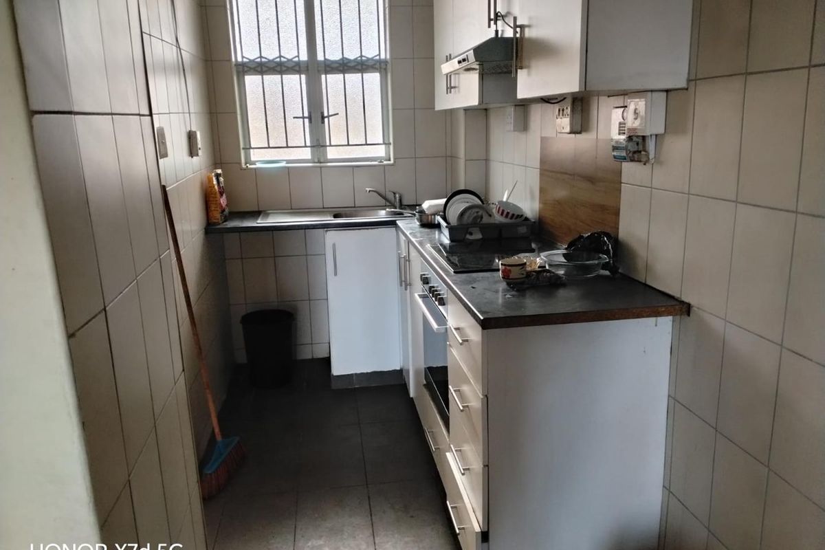 A compact galley-style kitchen featuring built-in cabinetry, an integrated oven and hob, tiled walls, and a window with security bars.