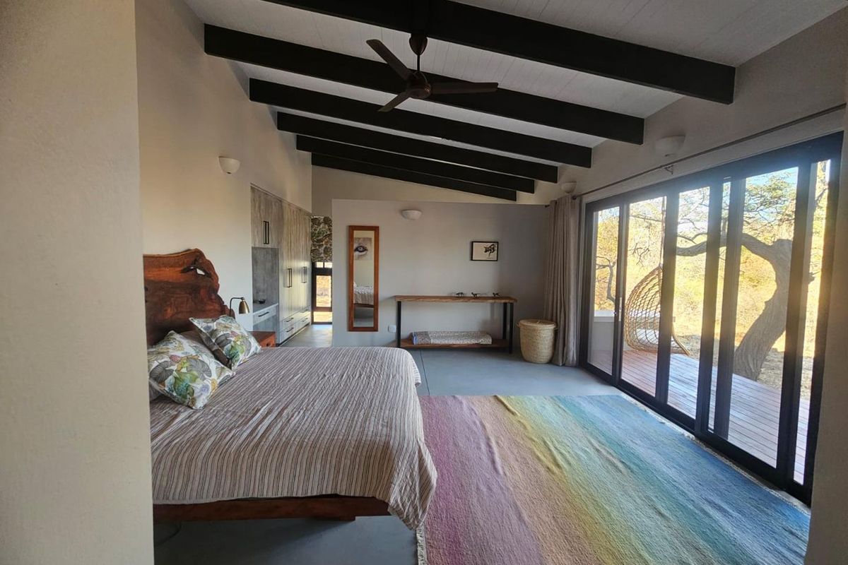 Spacious bedroom featuring exposed timber ceiling beams, polished concrete flooring, and large sliding glass doors providing direct access to an outdoor deck area.