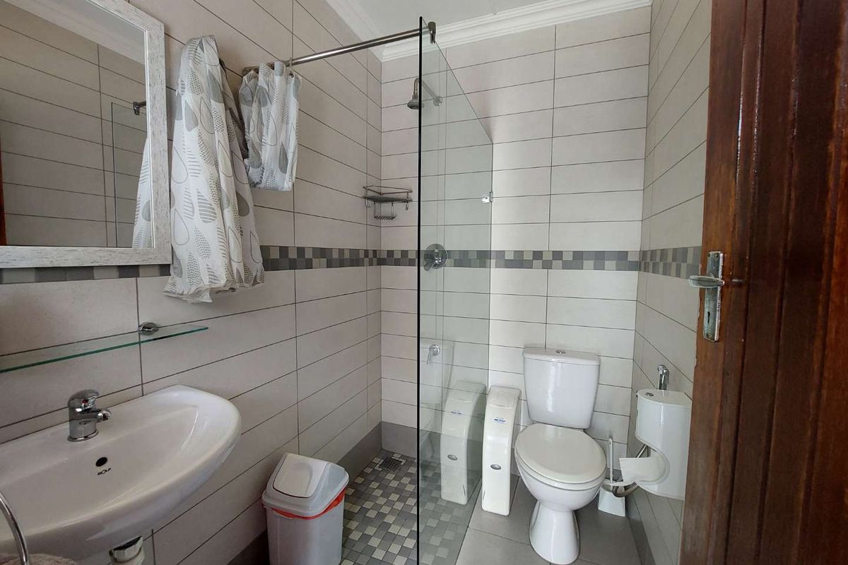 A compact office bathroom featuring a glass-enclosed shower, ceramic wall tiling, a pedestal basin, and a standard toilet.