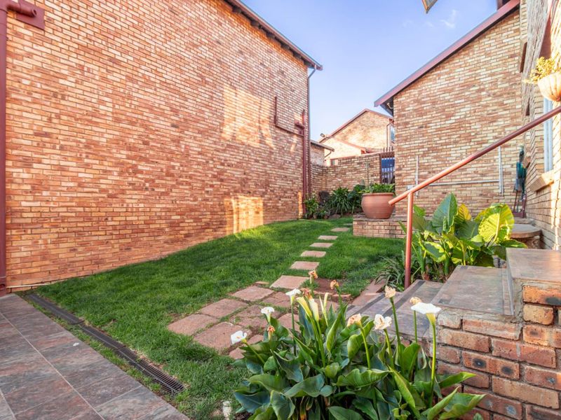 A well-maintained rear garden area featuring a brick facade, stepped tiled entrance, and a paved garden path leading through a lawn.