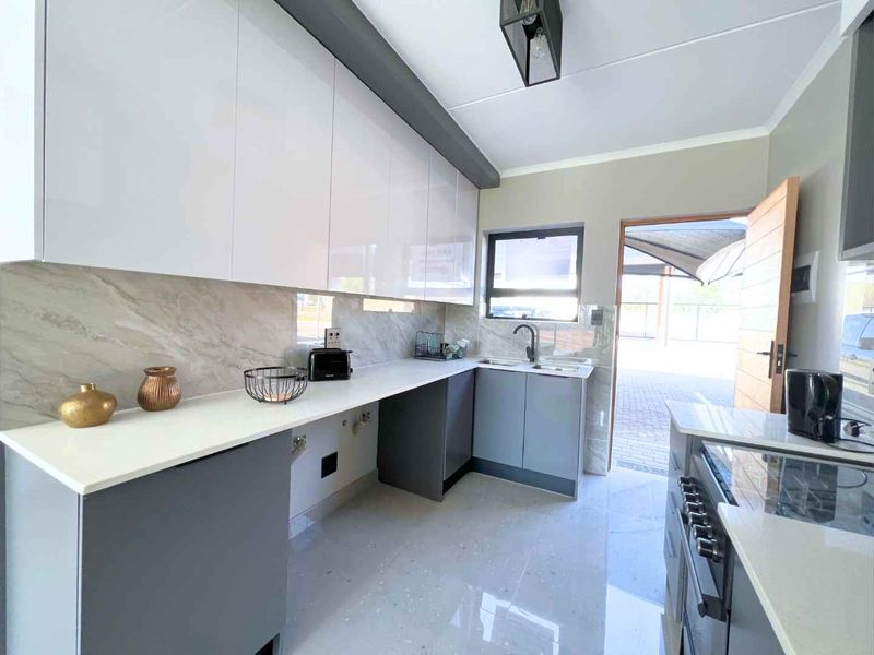 Modern kitchen featuring sleek white and grey cabinetry, stone countertops, a double sink, and a stylish stone splashback with direct outdoor access.