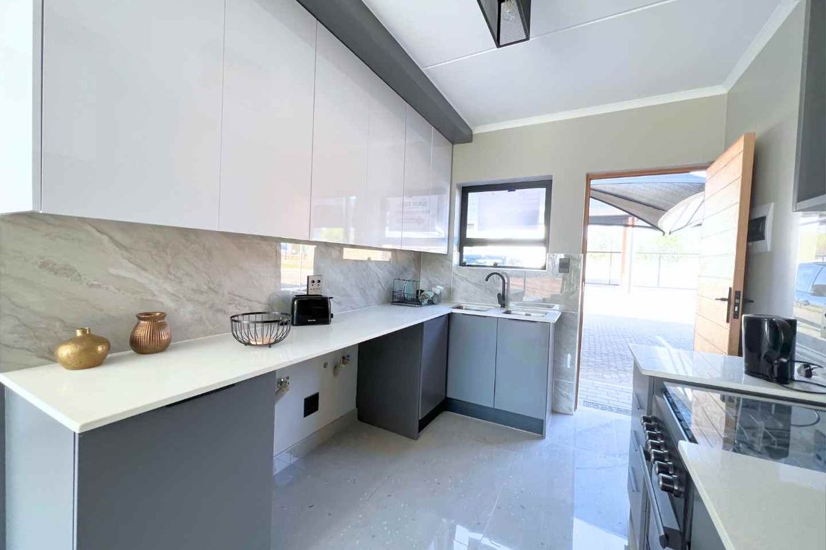 Modern kitchen featuring sleek white and grey cabinetry, stone countertops, a double sink, and a stylish stone splashback with direct outdoor access.