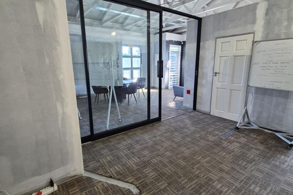 An office space featuring exposed roof trusses, a modern glass partition, and carpet tile flooring, currently in an unfinished state requiring cosmetic completion.
