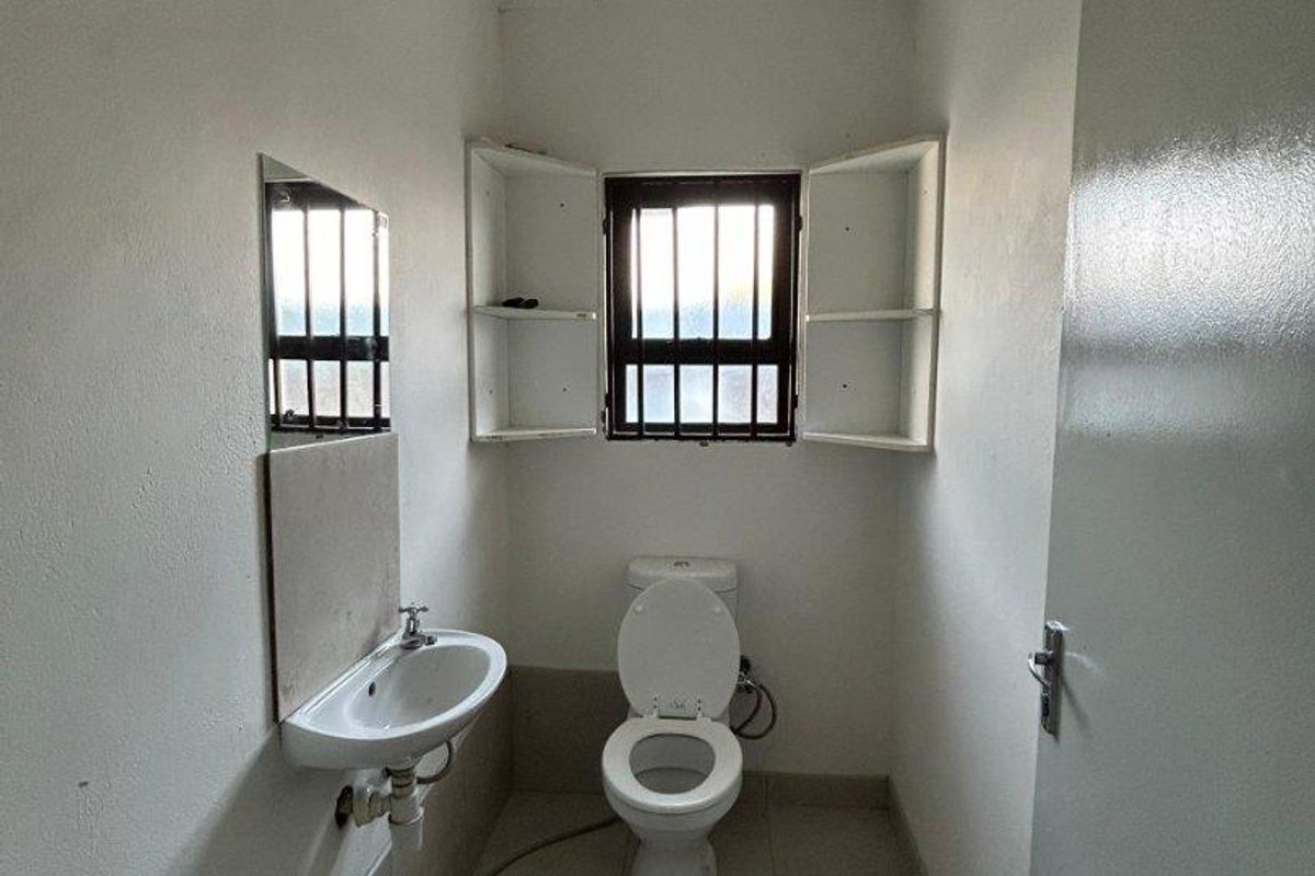 A compact bathroom featuring a wall-mounted basin, toilet, tiled flooring, and built-in shelving, with security bars fitted to the window.