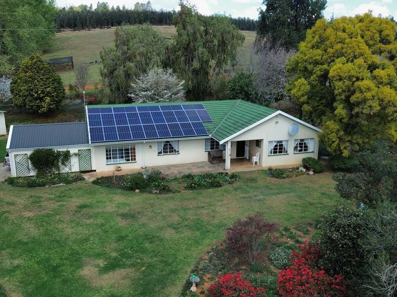 Aerial view of a farm property featuring a main residence with solar panels, a covered patio, and established garden surroundings.