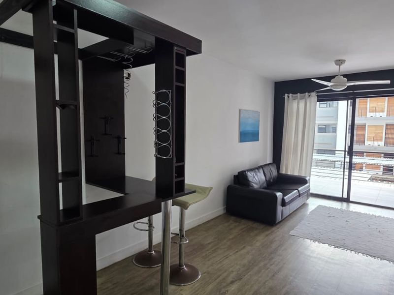 Open-plan living area featuring laminate flooring, a ceiling fan, and a built-in bar unit, with direct access to a balcony through sliding glass doors.