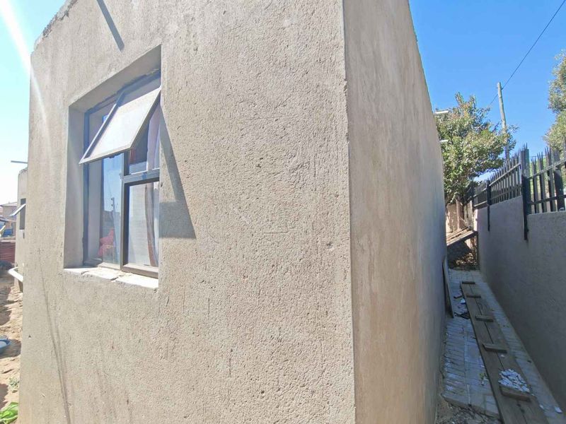 Side view of the property showing an unfinished plastered exterior wall, aluminium window frame, and a narrow side passage with metal perimeter fencing.
