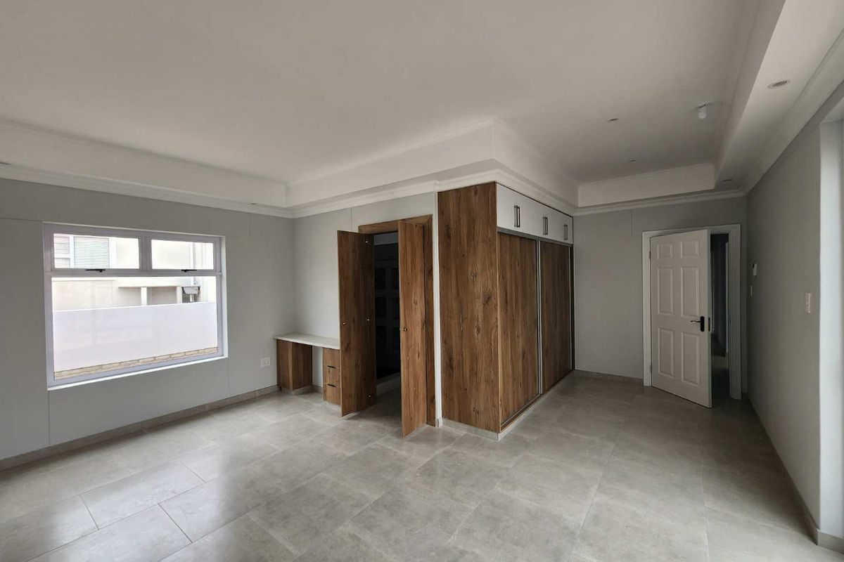 A spacious, modern bedroom featuring neutral tiled flooring, built-in wooden cabinetry, a dedicated desk area, and recessed ceiling lighting.
