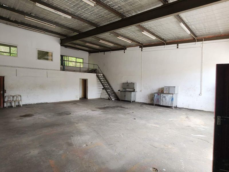 Spacious industrial utility area featuring high ceilings, exposed steel beams, a mezzanine level with staircase access, and concrete flooring suitable for workshop or storage use.