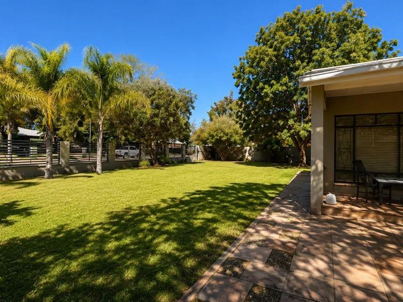 A spacious rear garden featuring a well-maintained lawn, mature trees, and a paved covered patio area enclosed by a perimeter wall and fence.